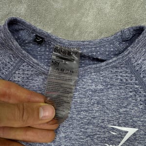 May include: A close-up of a light blue athletic shirt with the Gymshark logo in white. The shirt has a textured pattern and a tag with care instructions. The shirt is XS size.