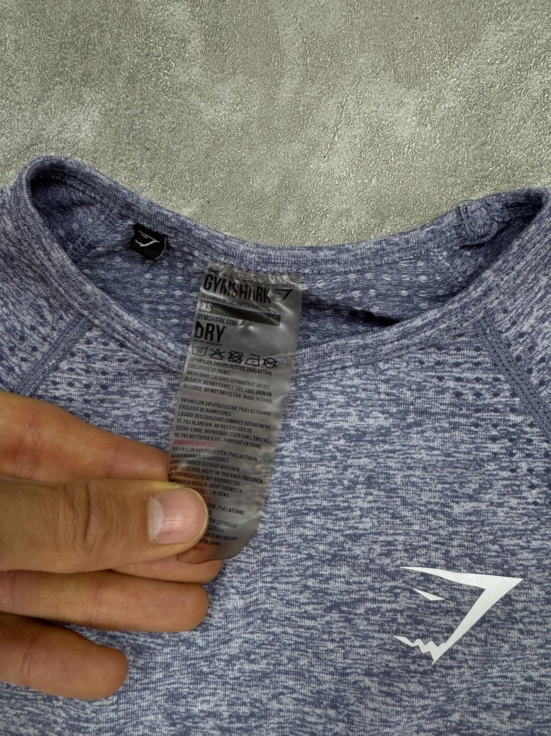 May include: A close-up of a light blue athletic shirt with the Gymshark logo in white. The shirt has a textured pattern and a tag with care instructions. The shirt is XS size.