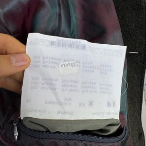 May include: Close-up of the inside of a dark green wool coat. The lining is a mix of burgundy and teal. A white tag with the number "0200000" is visible. A black zipper is on the bottom of the coat.