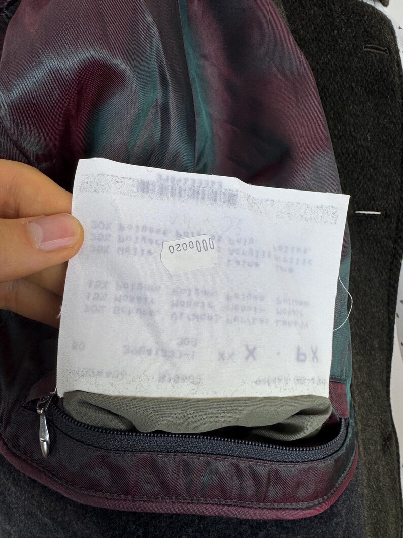 May include: Close-up of the inside of a dark green wool coat. The lining is a mix of burgundy and teal. A white tag with the number "0200000" is visible. A black zipper is on the bottom of the coat.