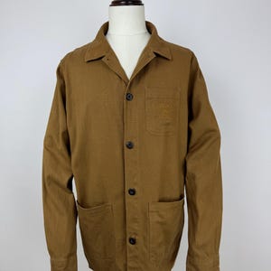 Könnte beinhalten: Braune Jacke mit Kragen, drei schwarzen Knöpfen und zwei Vordertaschen. Die Jacke hat lange Ärmel und eine kleine Tasche auf der Brust mit gesticktem Text. Die Jacke wird auf einer Schaufensterpuppe präsentiert.