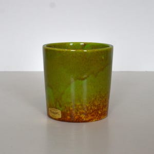 May include: A small, cylindrical ceramic cup with a glossy finish. The cup is primarily green, with a gradient of brown towards the bottom. A small, rectangular label is affixed to the side.