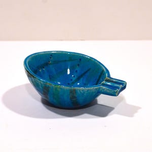 May include: A handcrafted, turquoise ceramic ashtray with a unique shape. The ashtray features a deep bowl with a built-in rest for cigarettes. The surface has a glossy finish with dark blue and green accents.