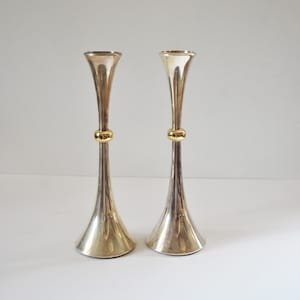 Puede incluir: Un par de candelabros altos y delgados de tono plateado con base y parte superior acampanadas. Cada candelabro está acentuado con un anillo dorado pulido colocado alrededor del centro del tallo, creando un contraste definido.