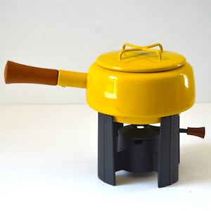 May include: A bright yellow fondue pot with a matching lid featuring a cross-shaped handle. The pot has a long, tapered wooden handle and rests on a dark gray metal stand that includes a small burner component with a wooden adjustment knob.