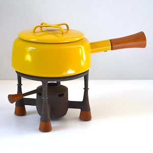 May include: A bright yellow enameled fondue pot with a matching lid and a long wooden handle. The pot rests on a dark gray metal stand featuring three legs with wooden feet and a central burner component with a small wooden adjustment lever.