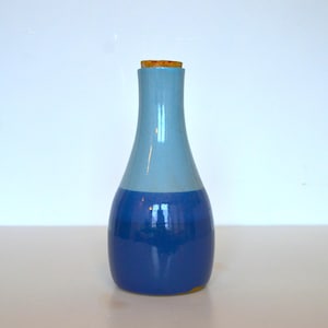 May include: A two-toned ceramic bottle with a cork stopper. The top half is light blue, and the bottom half is a darker blue. The bottle has a rounded base and a slender neck. The cork is a light brown color.