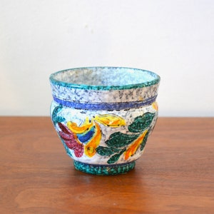 May include: A small, white ceramic planter with a green, blue, and yellow floral design. The planter has a speckled glaze and a rounded shape.