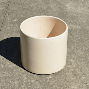 May include: A large, off-white ceramic planter with a smooth, rounded surface. The planter is sitting on a gray concrete surface.