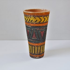 May include: A tall, cylindrical vase with a textured surface and a painted tribal design. The vase features bands of orange, yellow, green, and red geometric patterns. The vase is likely made of terracotta or a similar material.
