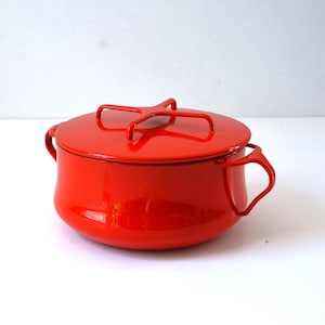 May include: A bright red enameled metal pot with a matching lid. The pot features two curved side handles and a cross-shaped handle on top of the lid. The exterior surface is smooth and glossy, showcasing a vibrant solid red color throughout.