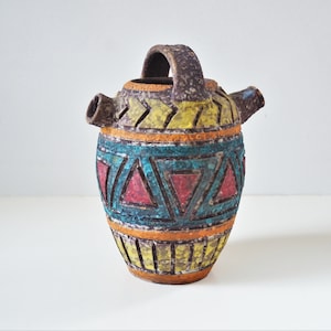 May include: A brown ceramic pitcher with a geometric design in yellow, orange, red, blue, and green. The pitcher has a handle and two spouts.