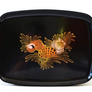 May include: A rectangular black serving tray with rounded corners featuring a central decorative motif of three seashells in shades of orange, gold, and white, surrounded by stylized golden seaweed or coral-like line patterns.