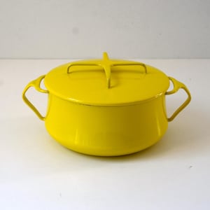 May include: A bright yellow enameled metal casserole dish with a matching lid. The pot features two curved side handles and a distinctive cross-shaped handle on top of the lid. The exterior has a smooth, glossy finish with a rounded base shape.