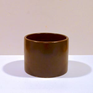 May include: A cylindrical planter with a smooth, dark brown glazed finish. The vessel features a simple, straight-sided design with a wide, open top and a flat base, presenting a uniform, solid color throughout the exterior surface of the object.