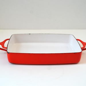 May include: A rectangular enameled cast iron baking dish featuring a vibrant red exterior and a smooth white interior. The cookware includes two integrated side handles and a black rim along the top edge, providing a durable and functional design.