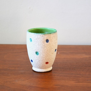 May include: A white textured ceramic vase with a green interior rim and colorful polka dots. The dots are blue, teal, and red. The vase is cylindrical with a slightly flared base and stands on a wooden surface.