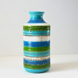 May include: A cylindrical ceramic vase featuring horizontal bands of blue, green, and white. The surface is decorated with intricate, repeating geometric patterns and textured carvings that wrap around the body, transitioning to a smooth blue neck.