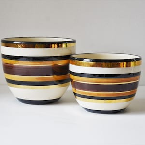 May include: Two ceramic planters with a striped design. The larger planter is white with black, brown, and gold stripes. The smaller planter is white with black, brown, and gold stripes.