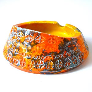 May include: Orange and yellow ceramic ashtray with a geometric design. The ashtray has a wide, shallow bowl and a raised rim.