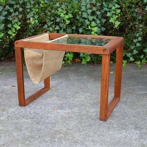 May include: A rectangular side table constructed from brown wood with a glass top insert. The table features a suspended beige fabric sling attached to the frame, designed for storage, with four legs forming a continuous rectangular base.