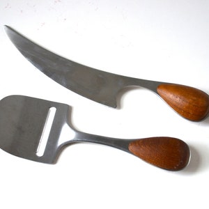 Puede incluir: Un cuchillo para queso de acero inoxidable con hoja curva y una cortadora de queso con cabeza plana ranurada. Ambos utensilios cuentan con mangos de madera lisos y redondeados en un tono marrón cálido y acabado metálico pulido.