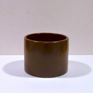 May include: A cylindrical, brown ceramic pot with a glossy finish. The pot has a wide opening and a smooth, rounded exterior. The interior is a darker shade of brown. The pot is likely used for plants or as a decorative item.