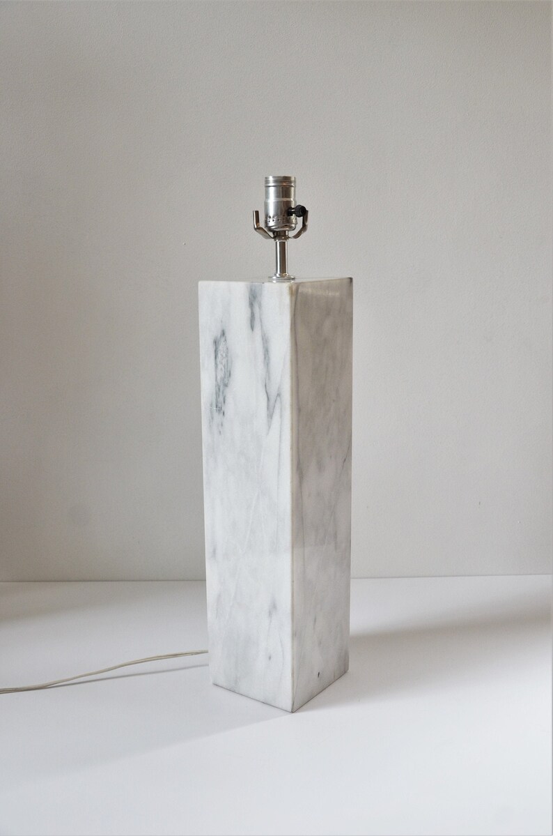 Puede incluir: Una l&aacute;mpara de mesa de m&aacute;rmol blanco con una pantalla plateada. La l&aacute;mpara est&aacute; sobre una superficie blanca.