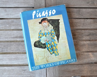 May include: A blue book with the title "Picasso: The Works of Picasso" featuring a painting of a young person wearing a blue and yellow harlequin costume sitting on a chair.