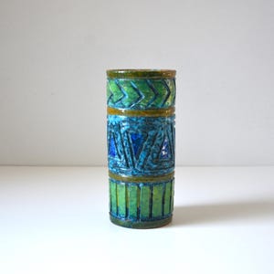 May include: A cylindrical ceramic vase with a geometric pattern in shades of blue, green, and yellow. The vase has a textured surface and a slightly flared opening.