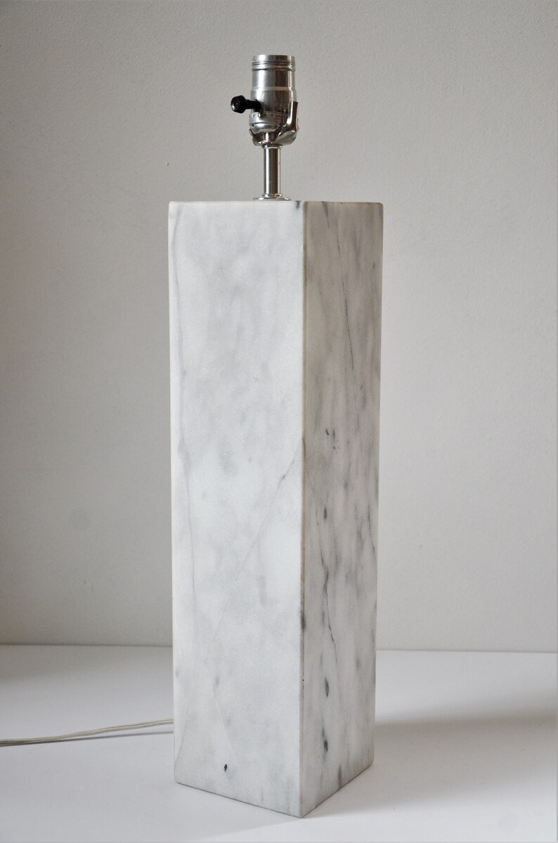 Puede incluir: Una base de l&aacute;mpara de m&aacute;rmol blanco con una toma de l&aacute;mpara plateada y un interruptor negro. La base es rectangular y tiene un acabado liso y pulido.