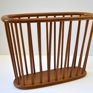 Puede incluir: Un revistero de madera con forma ovalada redondeada y listones verticales. El revistero es de madera marrón claro.