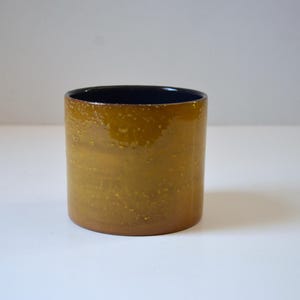 May include: A cylindrical ceramic mug with a glossy, textured finish. The exterior is a warm, golden-brown color, while the interior is a deep navy blue. The mug is a simple, yet elegant design, suitable for beverages.