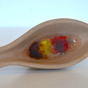 May include: A tan, spoon-shaped ceramic dish with a clear, oval-shaped well. The well contains a mix of colorful, abstract elements in red, yellow, and brown. The dish has a speckled texture.