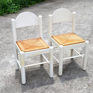 Puede incluir: Un par de sillas con estructuras de madera pintadas de blanco, respaldos redondeados y asientos tejidos de fibra natural en color tostado. Cada silla cuenta con cuatro patas cilíndricas unidas por barras de soporte horizontales.