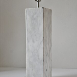 Puede incluir: Una l&aacute;mpara de mesa de m&aacute;rmol blanco con una pantalla plateada y un interruptor negro.