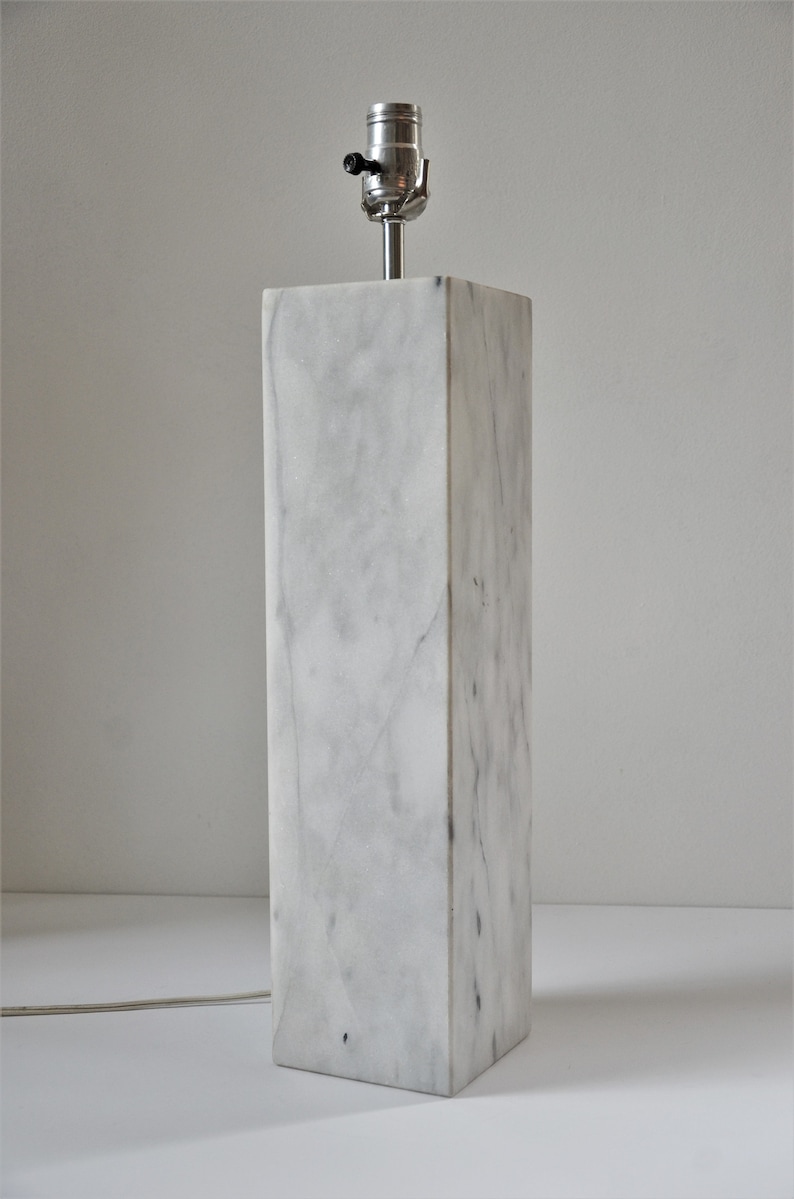 Puede incluir: Una l&aacute;mpara de mesa de m&aacute;rmol blanco con una pantalla plateada y un interruptor negro.