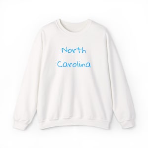 Puede incluir: Sudadera blanca de cuello redondo con las palabras "North Carolina" en azul claro. La sudadera de manga larga tiene un corte holgado, adecuada para el uso diario. El diseño es sencillo y limpio.