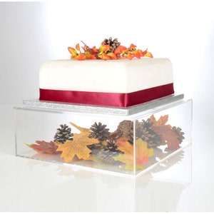 May include: A clear acrylic cake stand with a white cake decorated with orange and red flowers. The stand is filled with fall-themed decorations, including pinecones and leaves.