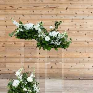 May include: Three clear acrylic pedestals of varying heights, each topped with a lush arrangement of white flowers and greenery.