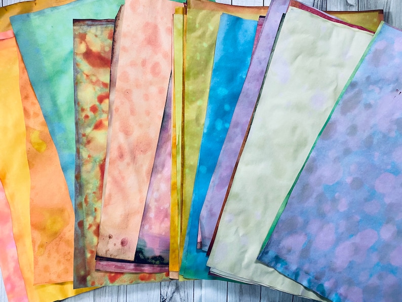 May include: A stack of colorful, hand-dyed paper sheets in various shades of blue, green, yellow, orange, and pink. The paper has a mottled, watercolor-like appearance.