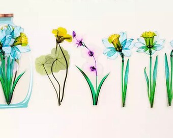 Può includere: Un set di sei adesivi floreali con narcisi blu e gialli e una singola foglia verde. Gli adesivi sono disposti in fila, con i narcisi in un vaso a sinistra e gli altri adesivi a destra.