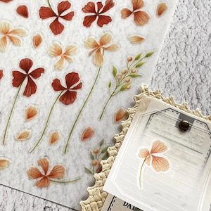 May include: A sheet of floral stickers featuring brown and peach-coloured flowers and petals. A decorative tag with a flower sticker is also visible. The stickers are on a white background.
