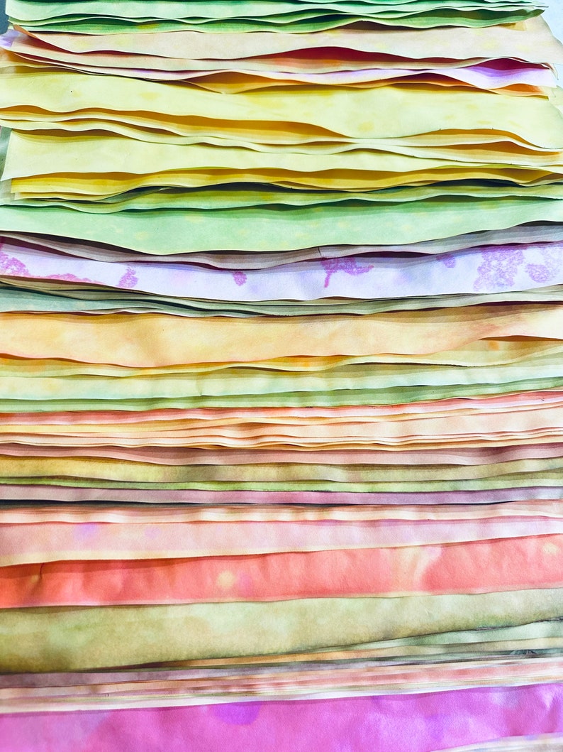 May include: A stack of colorful fabric strips in shades of yellow, green, orange, pink, and purple. The fabric is arranged in a layered pattern, creating a visually appealing and textured display.