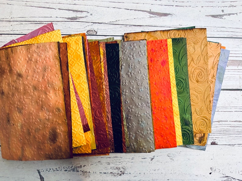 May include: A collection of textured paper sheets in various colors, including brown, yellow, orange, green, and blue. Each sheet has a unique embossed pattern, such as flowers and swirls. The papers are arranged in a fan shape.