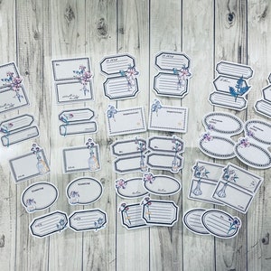May include: A collection of adhesive labels in various shapes including rectangles, ovals, and scalloped edges. Each label features blue and purple floral illustrations, glass bottle motifs, and lined sections for writing, all set against a white background.