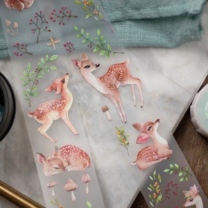 May include: A sheet of clear, translucent washi tape with a repeating pattern of watercolour illustrations of deer, mushrooms, and leaves. The deer are brown with white spots and are in various poses, including standing, lying down, and looking up.