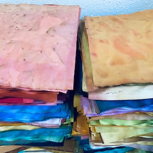 May include: Two stacks of handmade paper, one with a pink top sheet and the other with a brown top sheet. The stacks are made up of various colors of paper, including red, blue, green, yellow, and purple.