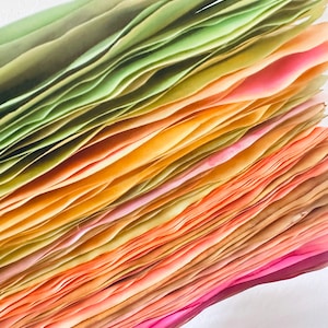 May include: A stack of colorful paper sheets, ranging from green to pink, with a gradient of yellow and orange in the middle. The paper is folded and layered, creating a textured and abstract pattern.