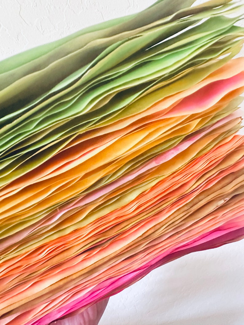 May include: A stack of colorful paper sheets, ranging from green to pink, with a gradient of yellow and orange in the middle. The paper is folded and layered, creating a textured and abstract pattern.